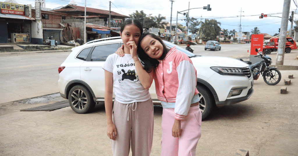 Chloe, also known as Ate Lottie, standing with her younger sister Jashel Kaye in a sweet outdoor sister photo near a white car.